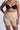 A person is wearing TITSY TUMMY TUCK SHORTS IN TAN with a black bra. Tattooed arms and red nail polish complement the look, set against a plain background. The image highlights their torso and upper legs.