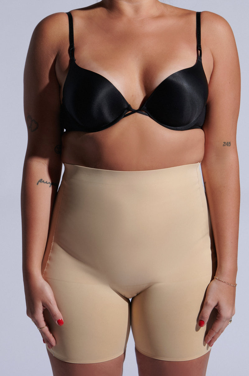 A person in a black bra and TITSY TUMMY TUCK SHORTS IN TAN with seamless hems stands against a light background, showing small arm tattoos and red-painted fingernails. 7