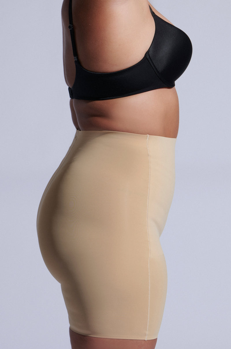 A person in profile showcases sleek TITSY TUMMY TUCK SHORTS IN TAN. These high-rise fit shapewear shorts have seamless hems that smooth the silhouette, set against a simple light gray background that highlights their elegance. 9