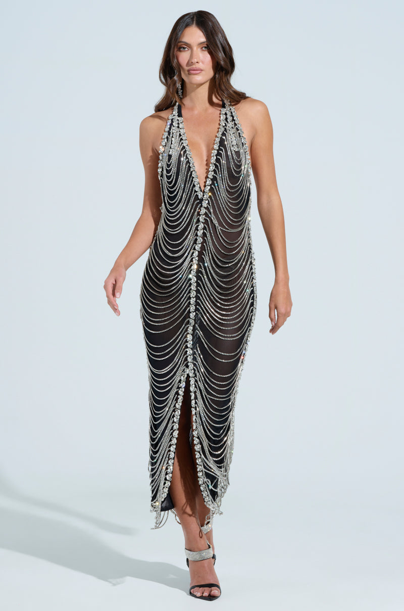 Someone walks forward, wearing the TALK IT OVER MAXI DRESS, a semi-sheer mesh maxi dress featuring intricate rhinestone detailing. This dress has a plunging neckline and a high slit, all set against a plain, light-colored background.