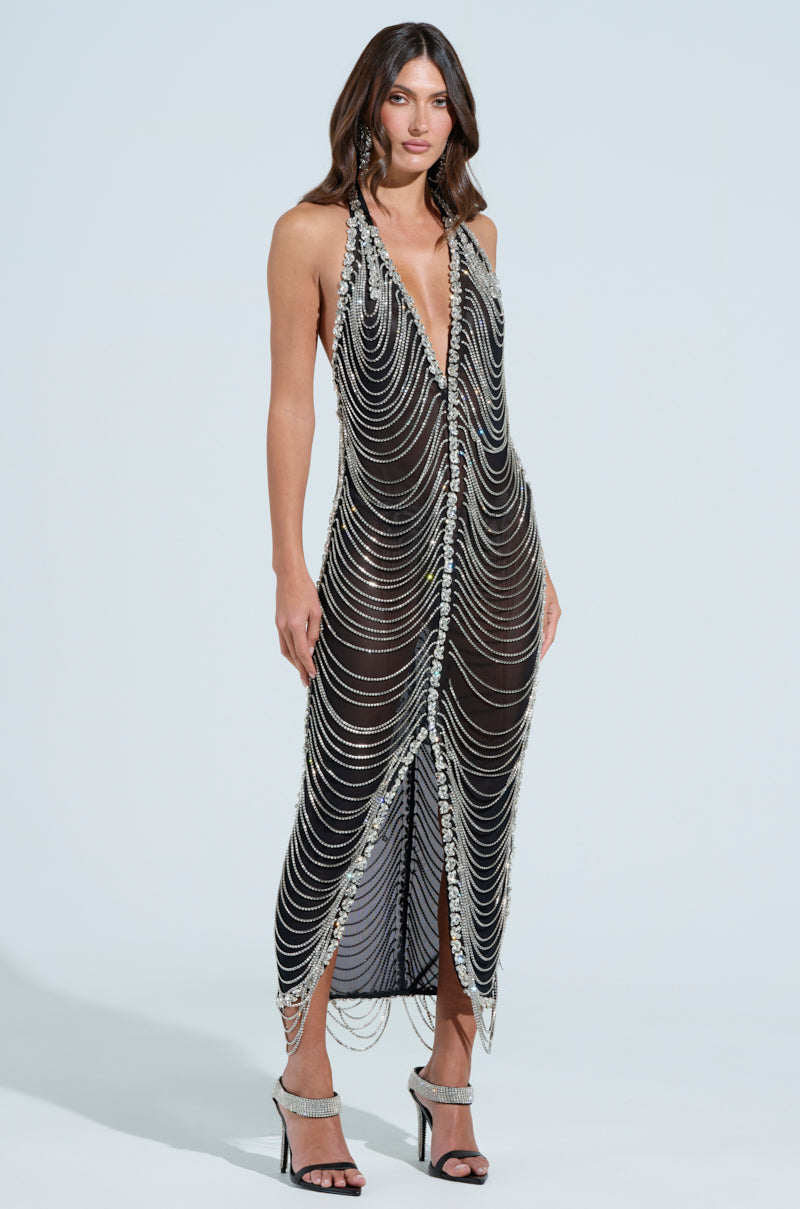 A woman in the TALK IT OVER MAXI DRESS, featuring a sleeveless, deep V-neck design with cascading silver chains and rhinestone detailing, stands in heeled sandals against a light blue background. The semi-sheer black mesh beautifully complements the metallic accents.
