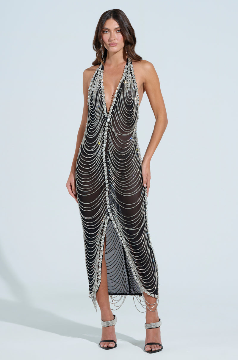 A woman poses elegantly against a white background in the TALK IT OVER MAXI DRESS, a floor-length black gown featuring a deep V-neckline accented with silver chains and rhinestones. She completes her look with sleeveless arms and strappy high-heeled sandals.