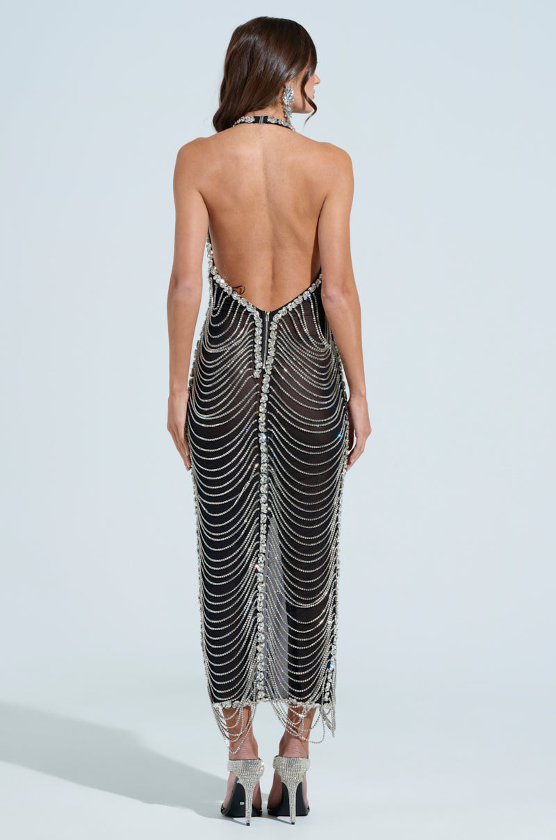 A woman in the TALK IT OVER MAXI DRESS, a beaded black backless design with intricate silver chain and rhinestone details, faces away. The semi-sheer mesh enhances its allure as she complements it with long earrings and high heels against a light blue backdrop.