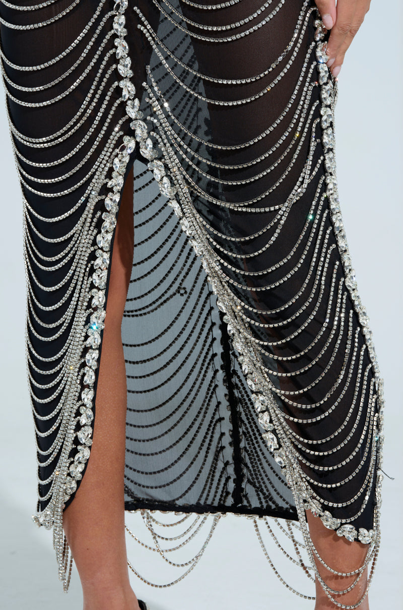 A close-up of someone wearing the TALK IT OVER MAXI DRESS, a black semi-sheer mesh design embellished with sparkling silver chains and rhinestones, featuring intricate cascading chain patterns and a leg-revealing slit.
