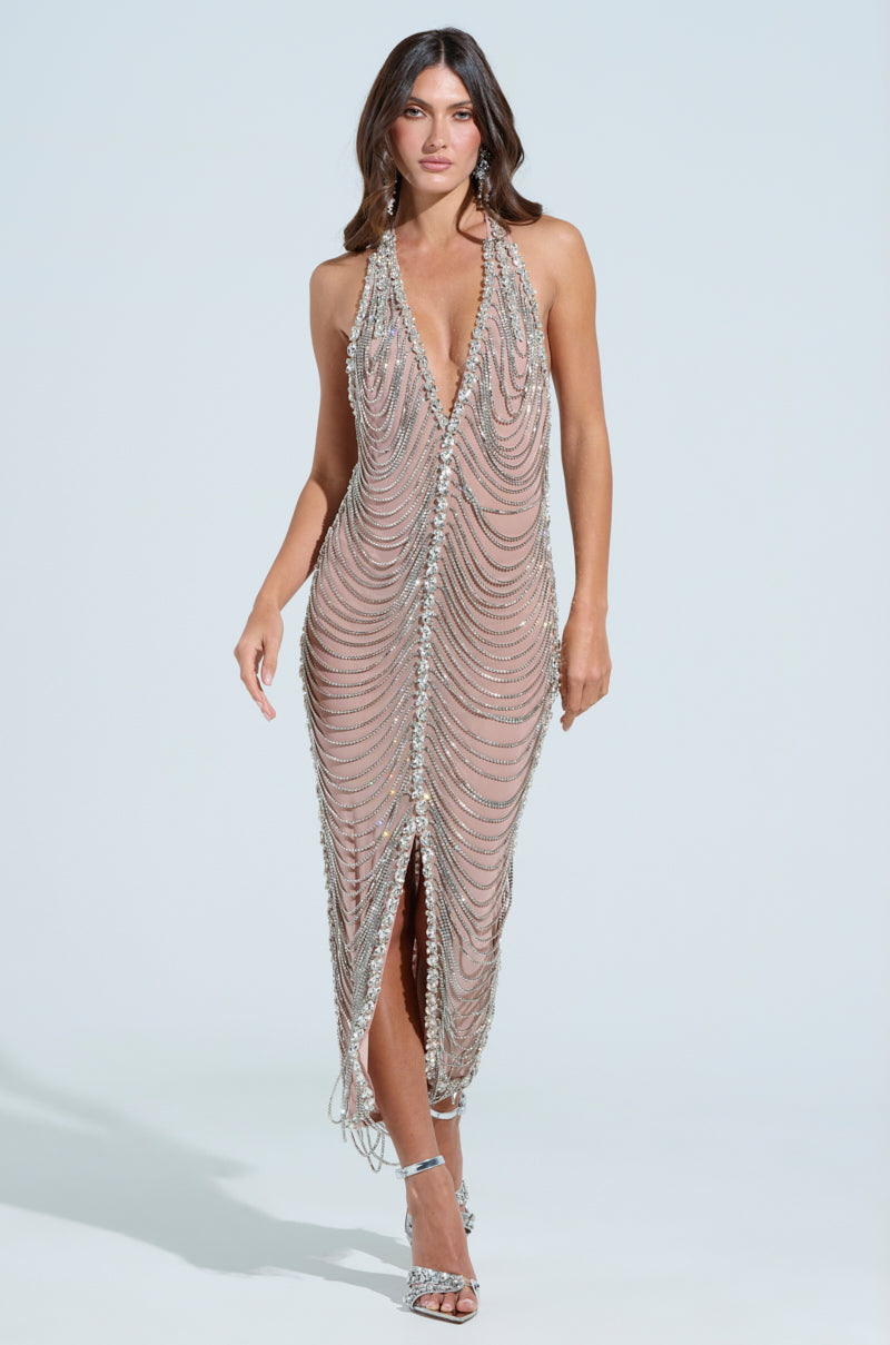 A woman confidently poses in a sparkling, silver-beaded DOMINATE CHAIN MAXI DRESS featuring a deep V-neck, high slit, and semi-sheer mesh with subtle rhinestone links. She pairs it with strappy silver heels and has long wavy hair, standing against a light blue background.
