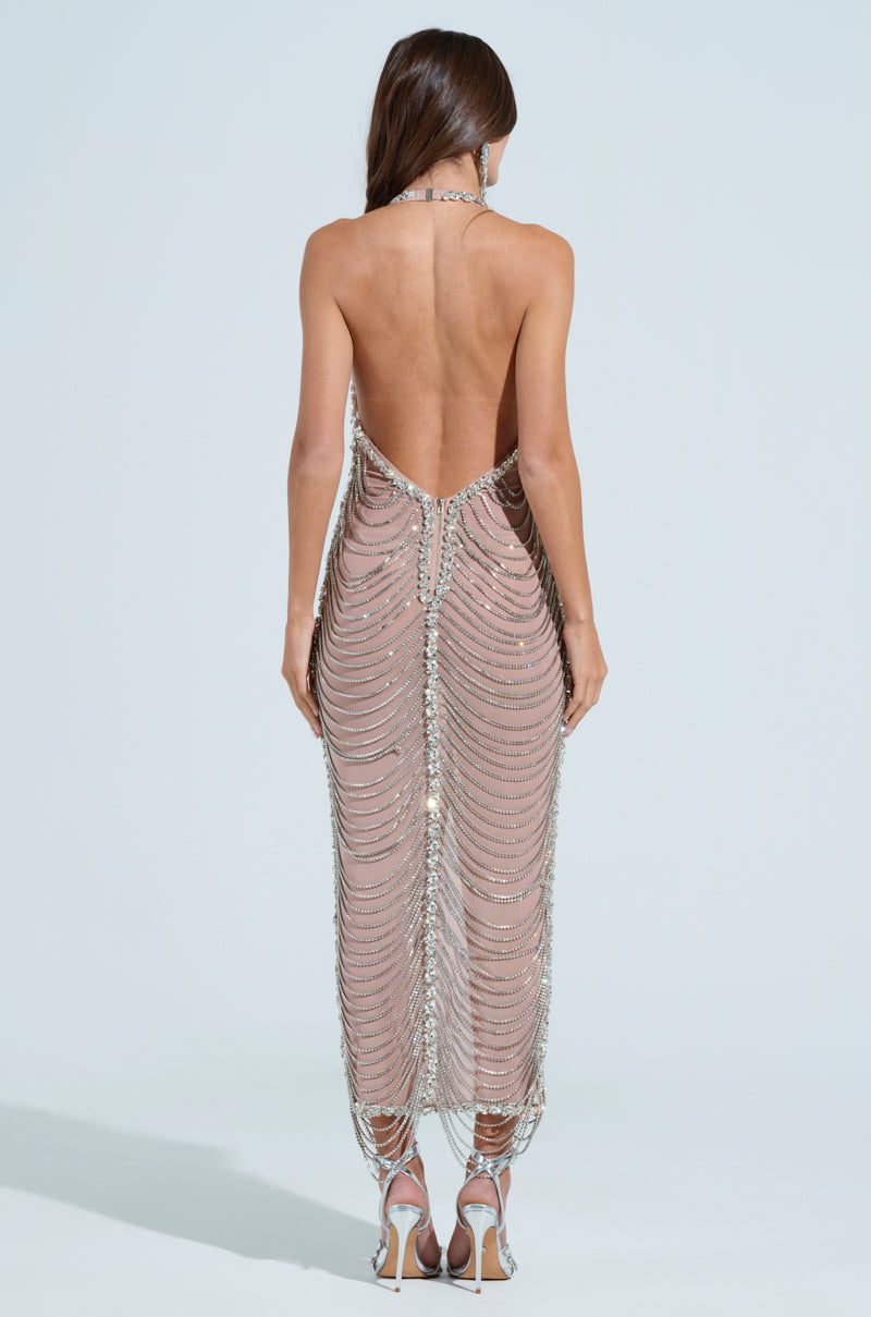 A woman stands with her back to the camera in a shimmering, backless DOMINATE CHAIN MAXI DRESS, decorated with silver beadwork and rhinestone links. The semi-sheer mesh gown reveals her high-heeled sandals against a light blue backdrop.