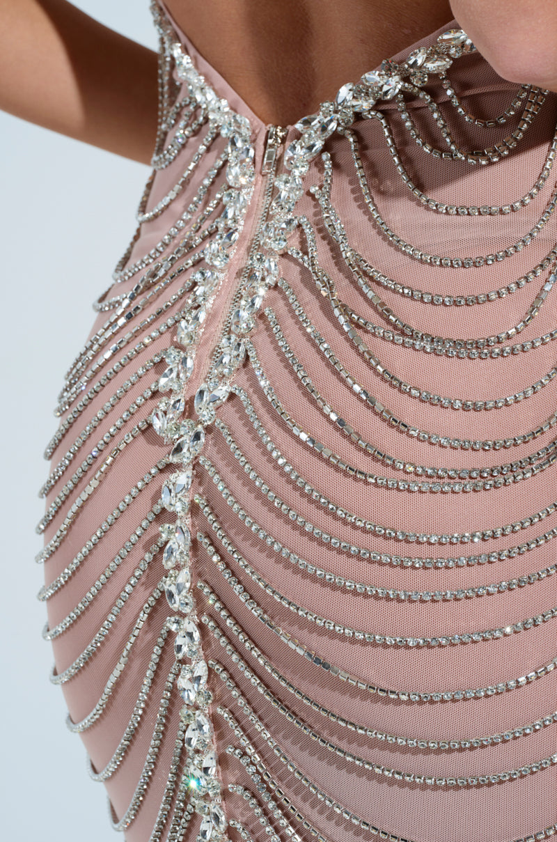 A person wearing a fitted, light pink DOMINATE CHAIN MAXI DRESS features silver rhinestone links in curved lines. The semi-sheer mesh adds allure, and the dress closes with a back zipper that highlights detailed beadwork.