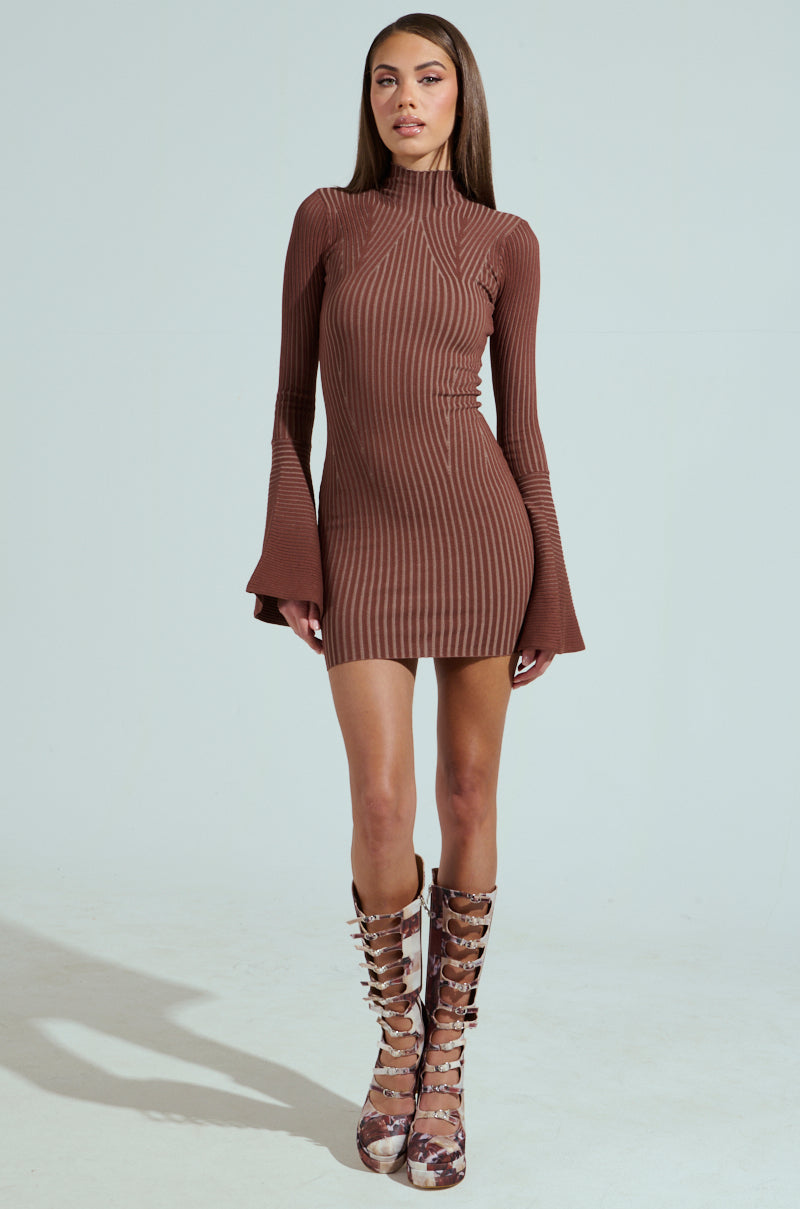 A woman exudes confidence in the ALL CAUGHT UP KNIT MINI DRESS, which features a curve-hugging ribbed knit and flared cuffs. She pairs it with knee-high, patterned studded boots against a plain, neutral background that beautifully highlights her outfit.