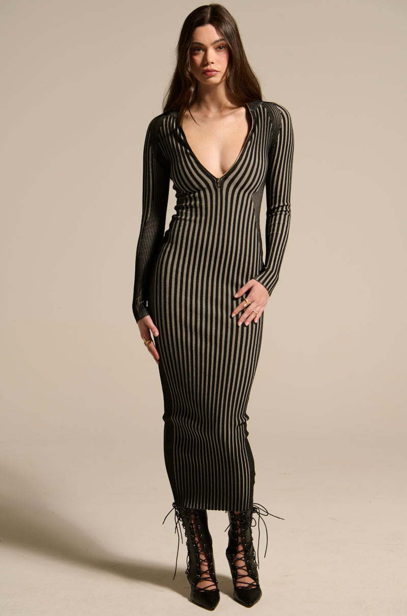 A woman with long brown hair wears the TAKE ME HOME KNIT MAXI DRESS, featuring a ribbed texture, black and beige stripes, deep V-neckline, and long sleeves. She stands facing forward in black lace-up boots against a neutral background.