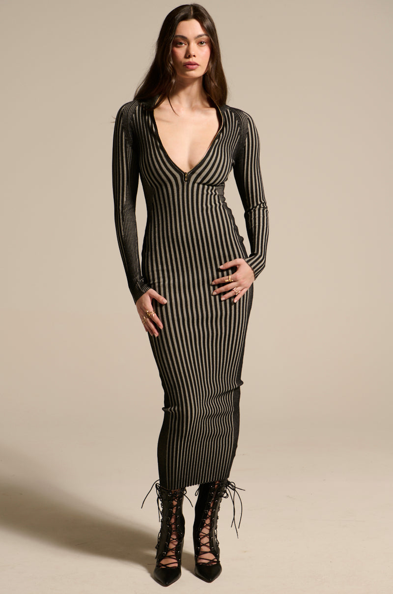 A woman with long hair wears the TAKE ME HOME KNIT MAXI DRESS, a fitted, long-sleeve black and beige knit maxi with vertical stripes and a deep V-neckline, styled with black lace-up boots against a plain background.