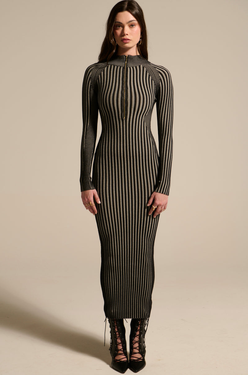 A woman faces forward wearing the TAKE ME HOME KNIT MAXI DRESS, featuring vertical black and beige stripes, a metallic gold zip front, long sleeves, and black lace-up shoes. Her brown hair is worn long.