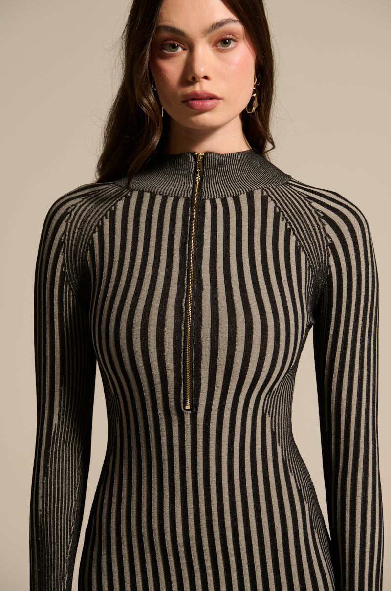 A woman wears the TAKE ME HOME KNIT MAXI DRESS, a fitted maxi with vertical black and gray stripes, a metallic gold front zip, and ribbed texture. She has long brown hair and stands against a plain light background.