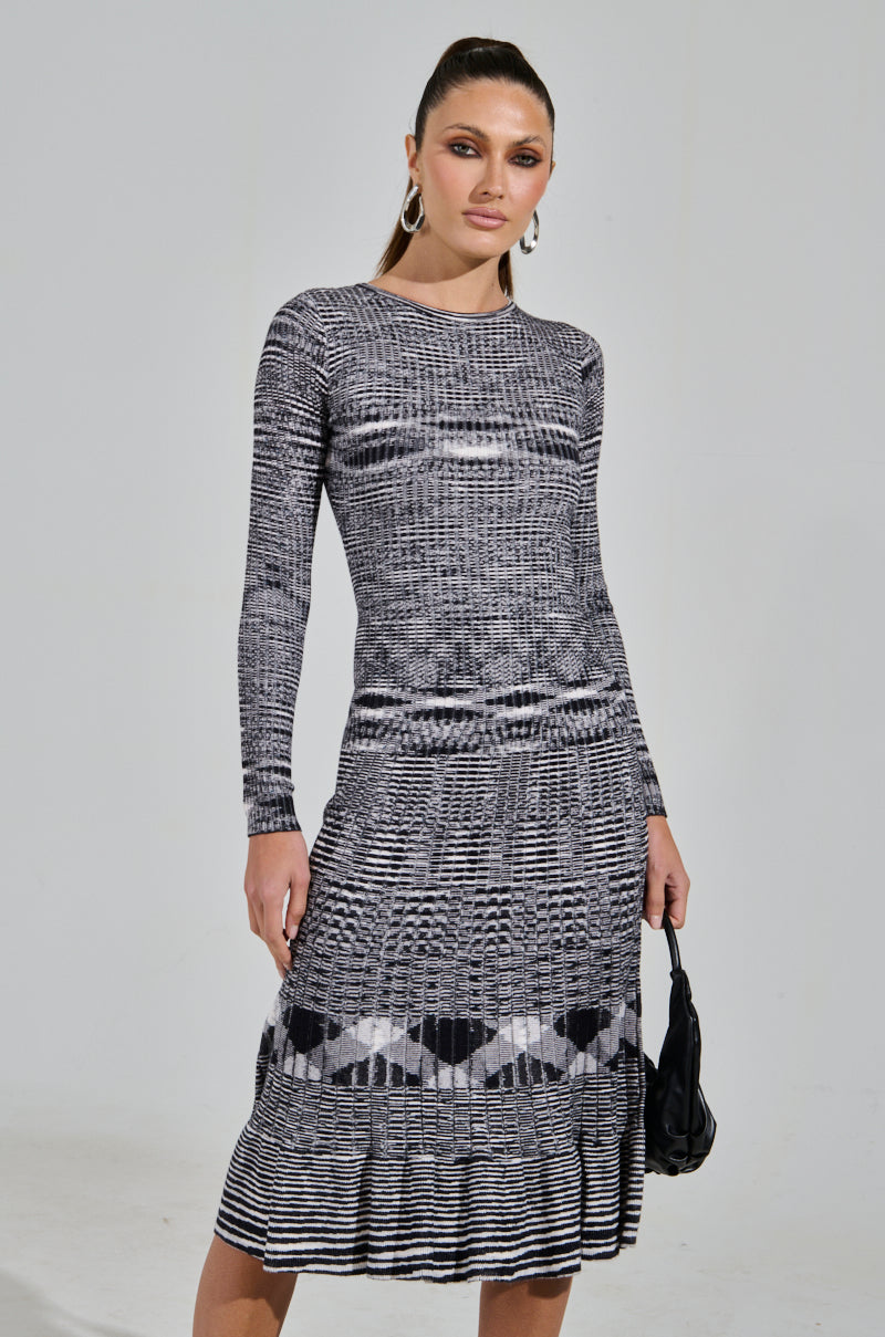 Wearing the SWERVIN KNIT MAXI DRESS, a person stands against a plain background. The black and white patterned, curve-hugging dress is complemented by hoop earrings and a small black handbag.