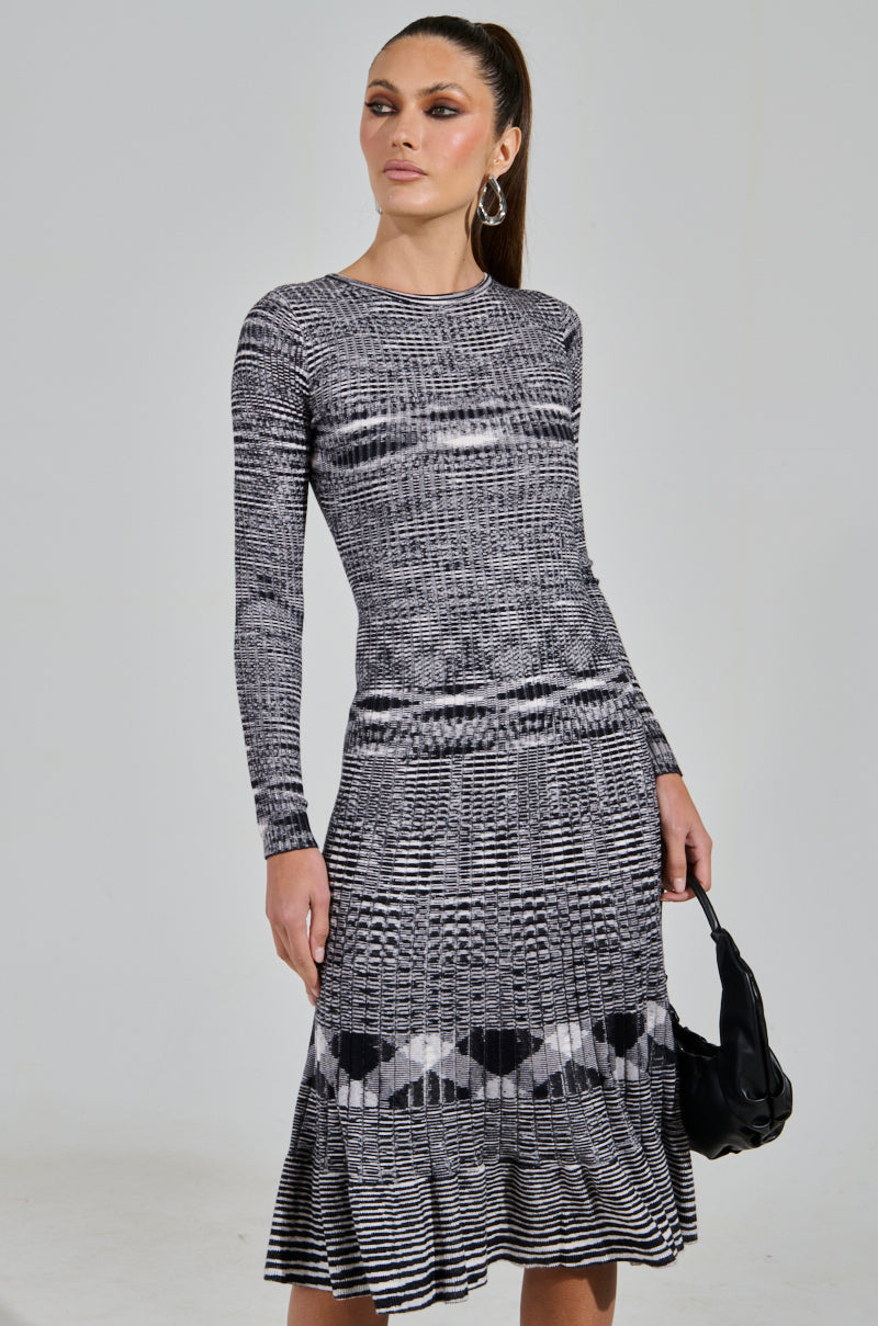 Wearing the SWERVIN KNIT MAXI DRESS, a person poses against a plain background. The long-sleeved, black-and-white patterned dress enhances their style with its curve-hugging silhouette. They hold a small black handbag, and their long hair is elegantly tied back.
