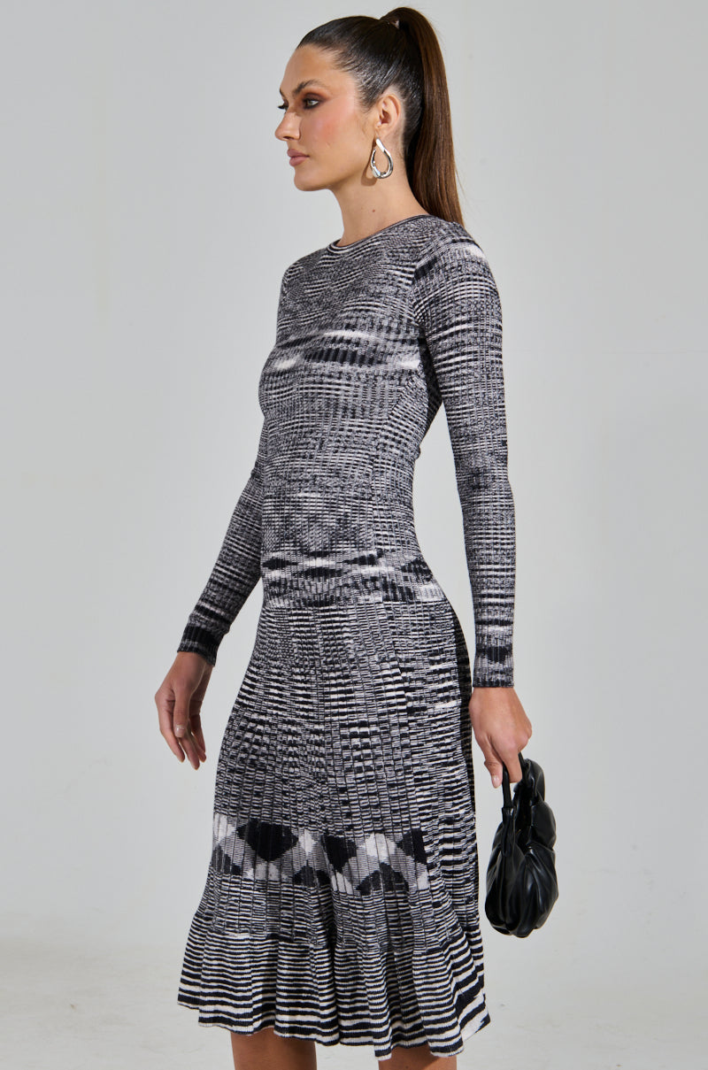 A woman poses gracefully in the SWERVIN KNIT MAXI DRESS, a long-sleeved black and white patterned knit maxi. She carries a small black handbag, large hoop earrings glinting under the light, her hair styled in a high ponytail against a plain backdrop.