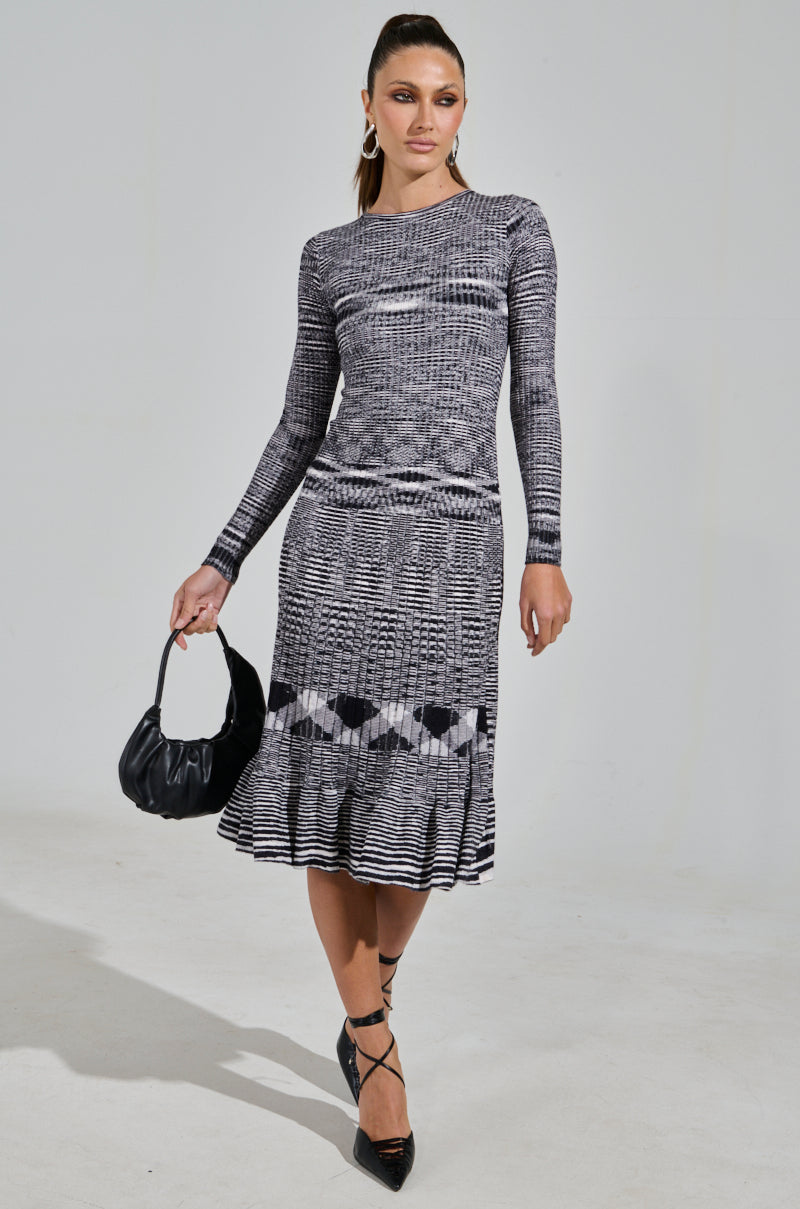A woman in the SWERVIN KNIT MAXI DRESS, featuring a black and white pattern and stretchy knit fabric for a curve-hugging silhouette, holds a small black handbag. She complements the look with black heels and hoop earrings, standing confidently against a plain background.