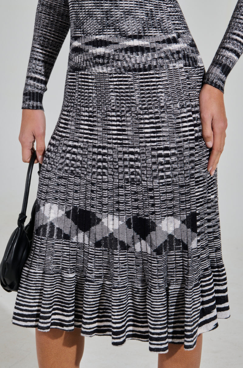 A person wearing the SWERVIN KNIT MAXI DRESS, a black and white geometric patterned maxi dress with long sleeves and a ruffled hem, holds a small black handbag. The stretchy knit fabric varies in texture and stripe width for a curve-hugging silhouette.