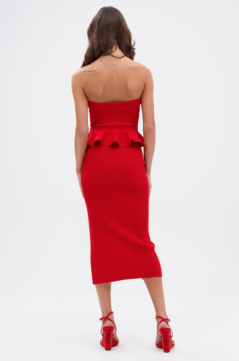 A woman with long brown hair stands with her back to the camera in the KIKI MIDI DRESS IN RED, a ribbed knit strapless midi dress with peplum detail, paired with red high-heeled sandals against a white background.