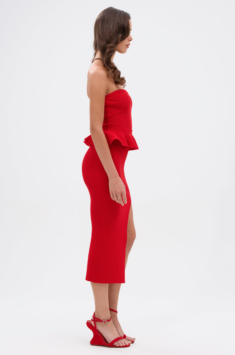 Woman in profile against a white background wears the KIKI MIDI DRESS IN RED, featuring a strapless silhouette, peplum waist detail, and side slit, paired with red wedge sandals.