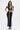A woman poses with one hand on her hip in the LUCKY NUMBER 7 MAXI DRESS, a fitted black mesh maxi with varsity-style bold white trim, paired with a small white bag and strappy black heels against a plain white background.