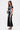 A woman with long brown hair wears the LUCKY NUMBER 7 MAXI DRESS—a varsity-style black mesh maxi dress with white stripes and a number 7 on the chest. She holds a black and white purse and pairs the look with black open-toe heels.