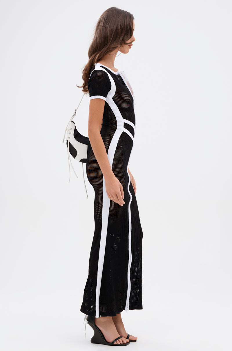 A woman stands sideways in the LUCKY NUMBER 7 MAXI DRESS—a varsity-style black sheer mesh dress with bold white stripes—carrying a small black-and-white handbag and wearing black wedge sandals against a plain white background.