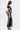 A woman stands sideways in the LUCKY NUMBER 7 MAXI DRESS—a varsity-style black sheer mesh dress with bold white stripes—carrying a small black-and-white handbag and wearing black wedge sandals against a plain white background.