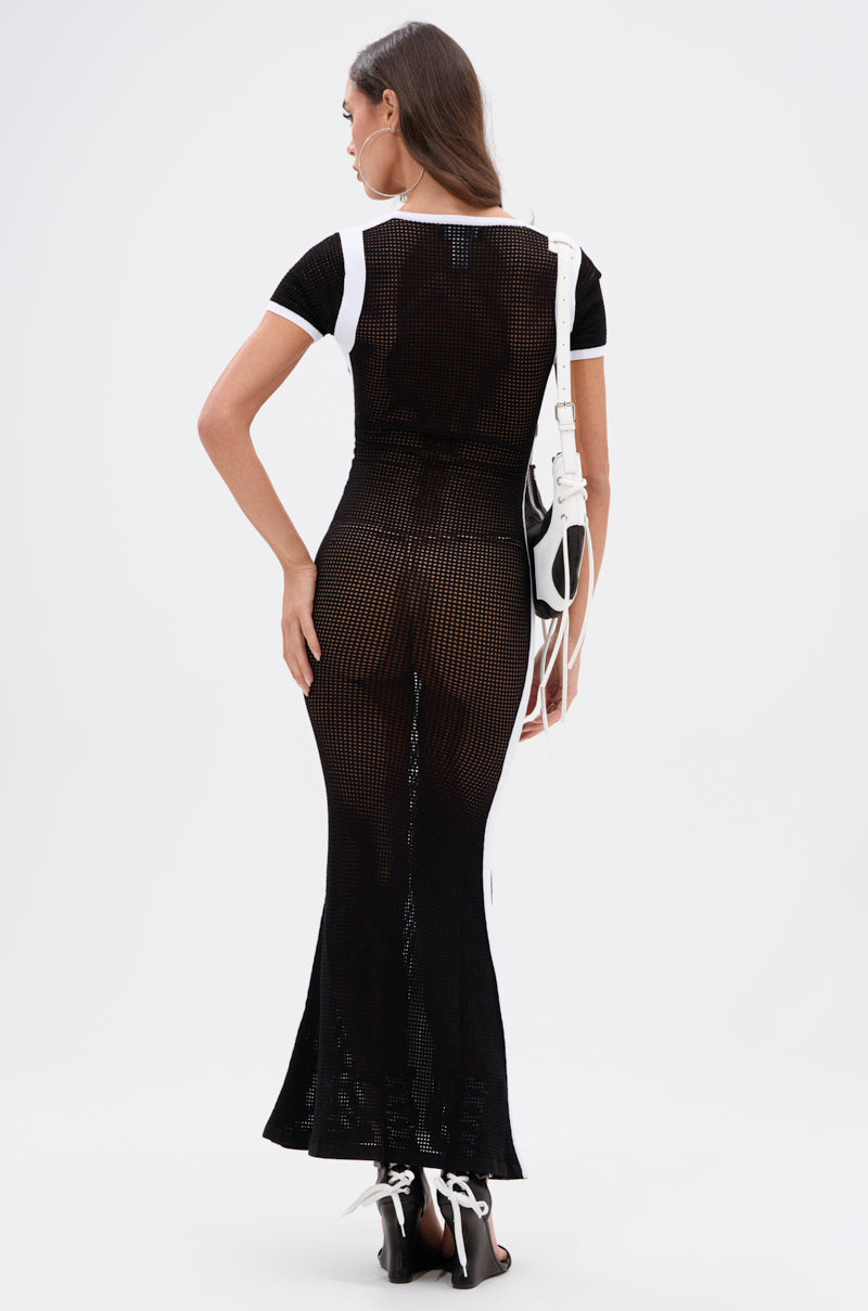 A woman faces away wearing the LUCKY NUMBER 7 MAXI DRESS, a varsity-style black mesh maxi with white trim that reveals black underwear. She carries a white purse and pairs the look with black and white high-heeled sandals.