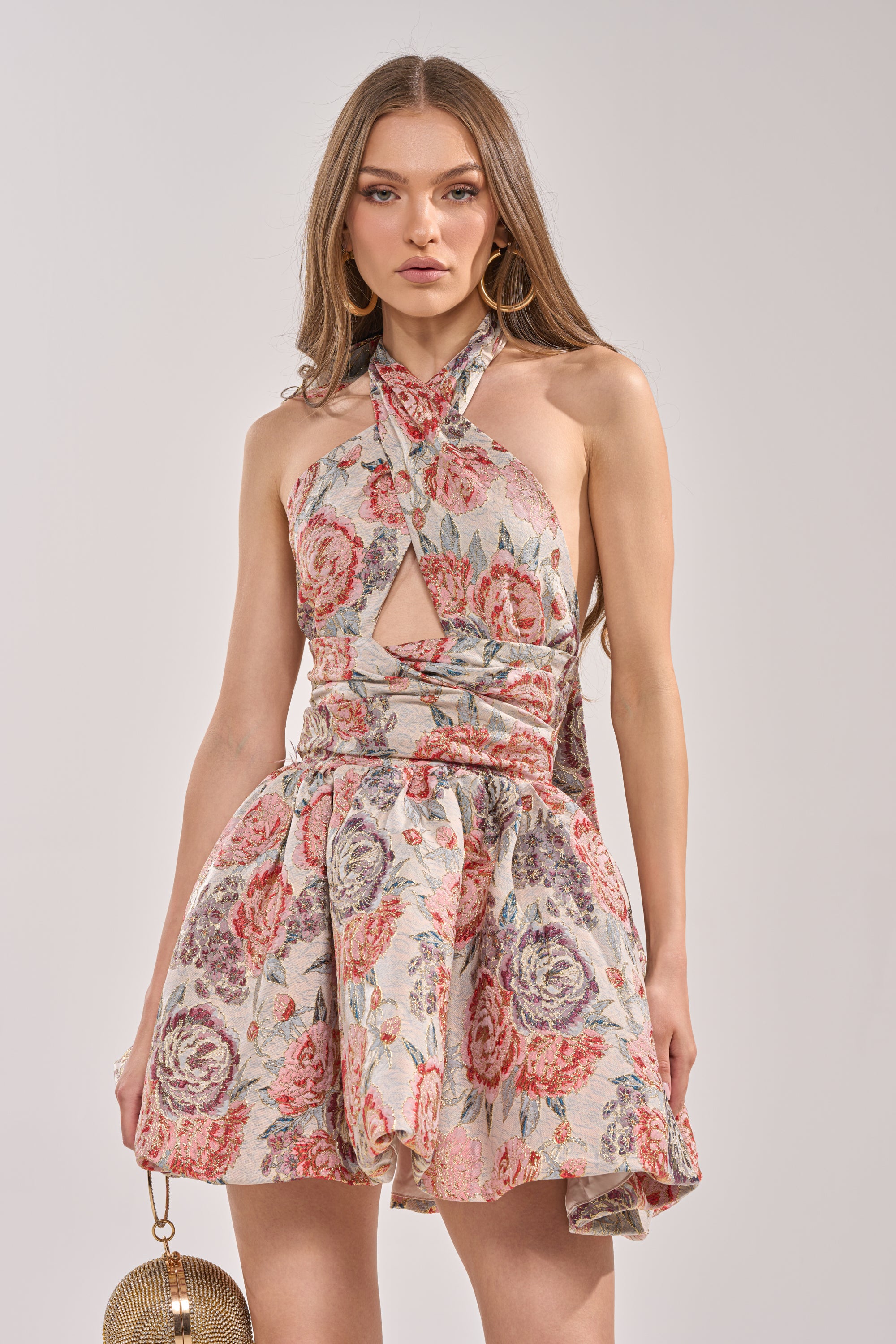 A woman in the PRINT OF THE SEASON BUBBLE MINI DRESS IN BROCADE—sleeveless, halter-neck, with a chest cutout—stands against a plain background, holding a round gold handbag. 1