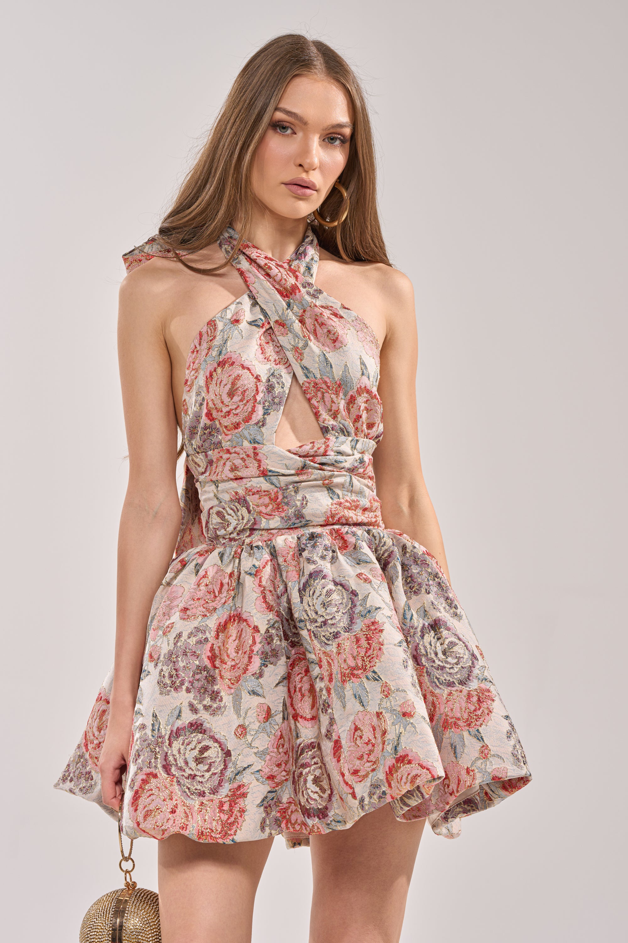 A woman wears the PRINT OF THE SEASON BUBBLE MINI DRESS IN BROCADE—a sleeveless, halter-neck floral brocade mini dress with pink, red, and gray tones, a front cut-out, and flared skirt—posing with a gold purse against a plain background. 3