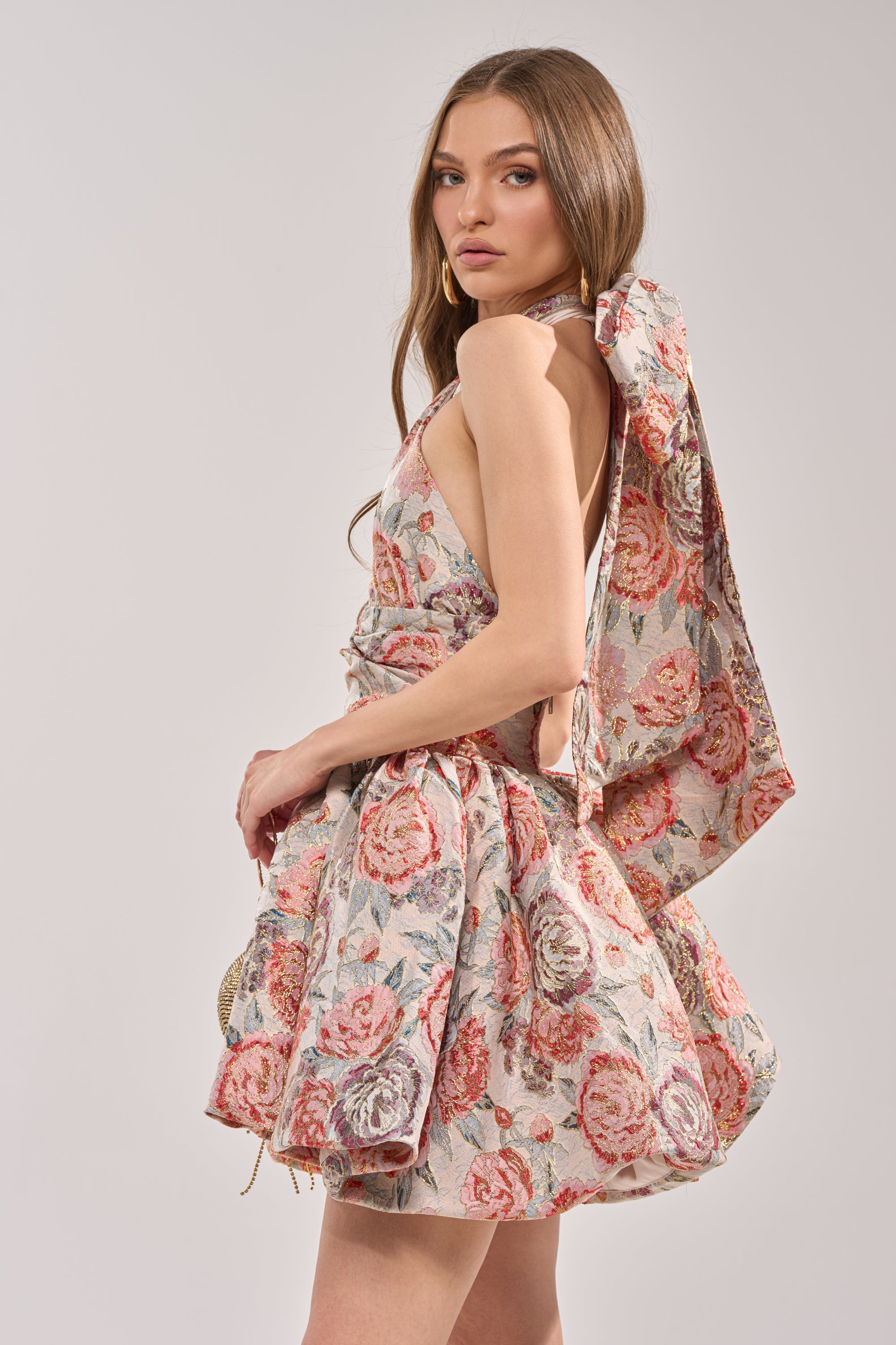A woman poses sideways against a plain background, looking over her shoulder in the PRINT OF THE SEASON BUBBLE MINI DRESS IN BROCADE, a sleeveless floral mini dress featuring a large bow at the back. 4