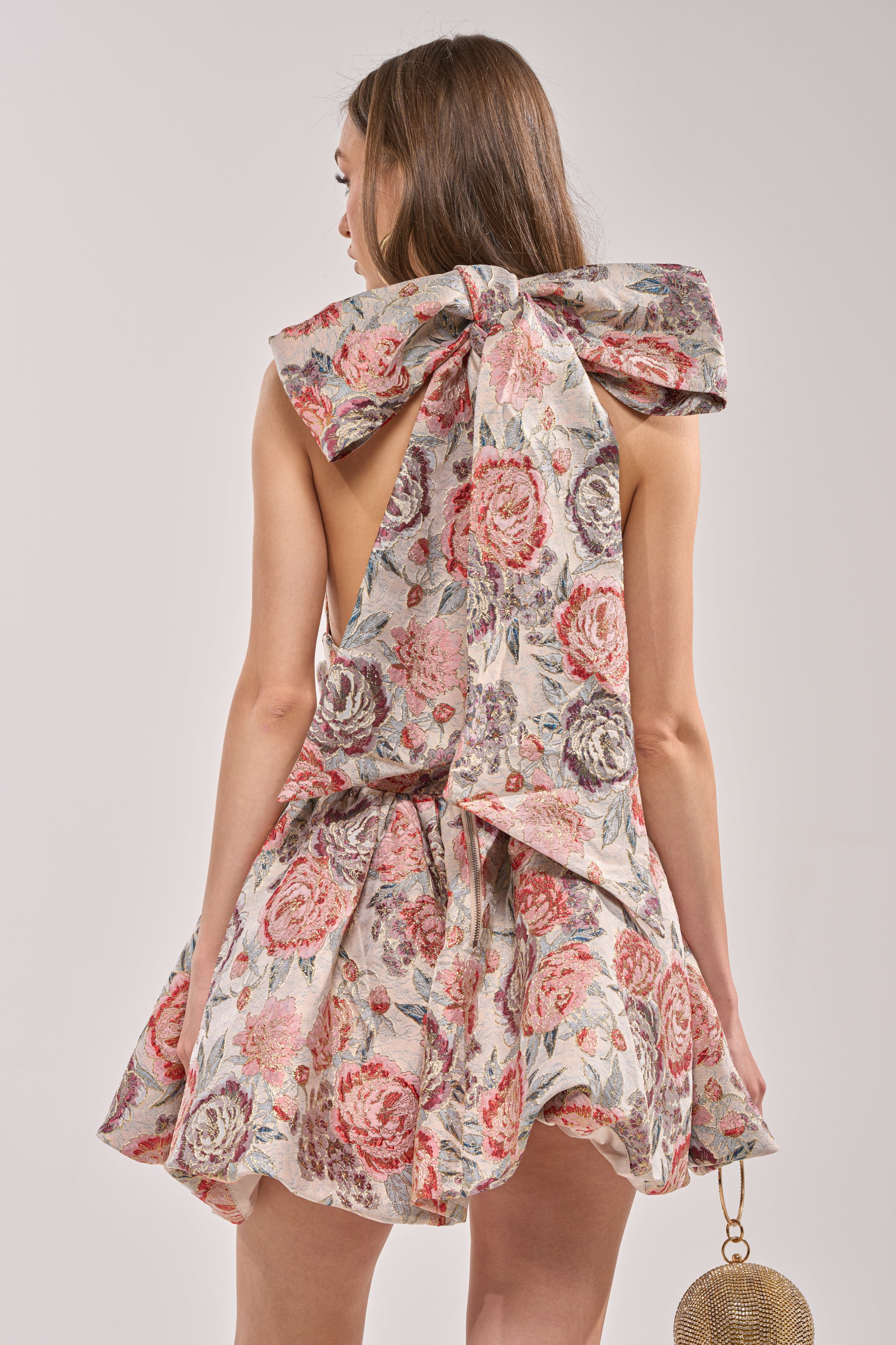 A woman in the PRINT OF THE SEASON BUBBLE MINI DRESS IN BROCADE, a sleeveless mini with pink, red, and green floral patterns and a large back bow, stands facing away holding a small round gold bag. 5
