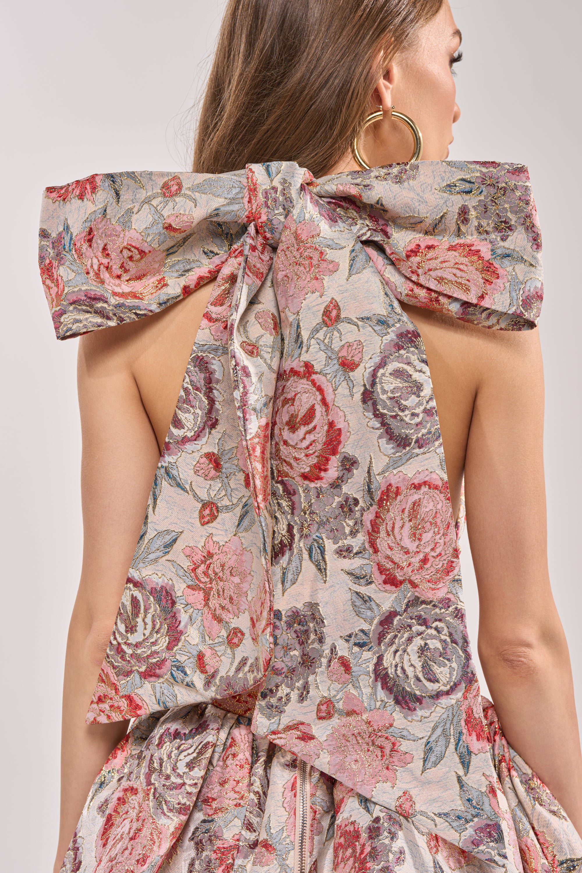 A woman with long brown hair wears the PRINT OF THE SEASON BUBBLE MINI DRESS IN BROCADE, featuring a pink, red, and green rose pattern and a large bow on the back. She completes her party look with bold hoop earrings. 8