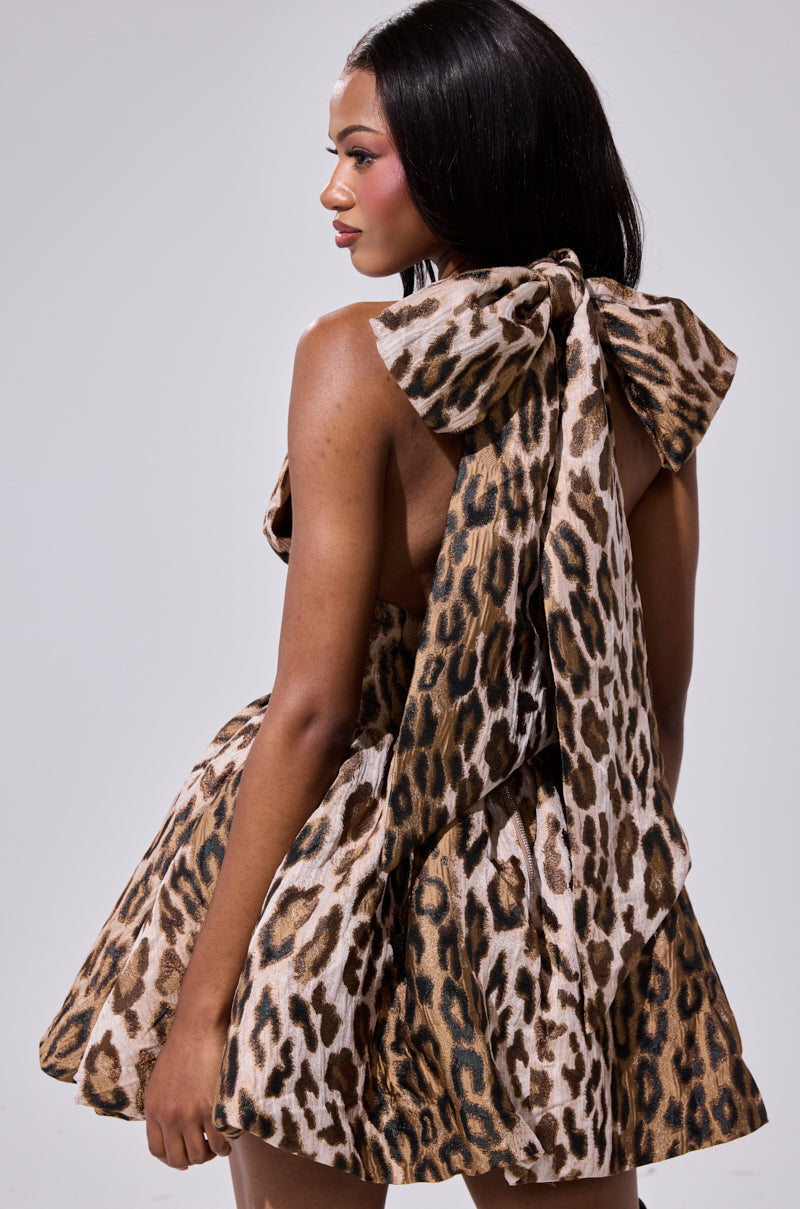 A woman models the PRINT OF THE SEASON BUBBLE MINI DRESS—featuring leopard print, a halter neckline, and a statement bow at the back—as she stands against a plain background, gazing to the side with her hand by her side.
