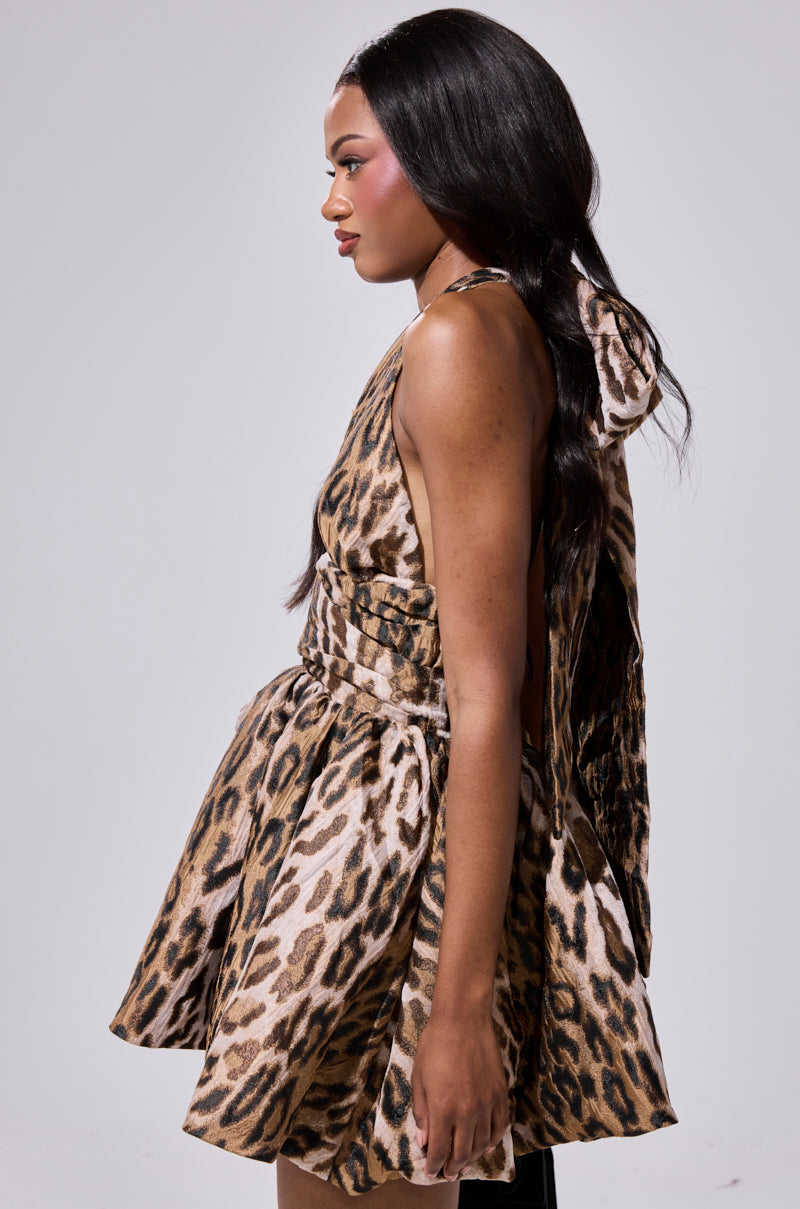 A woman stands in profile wearing the PRINT OF THE SEASON BUBBLE MINI DRESS, featuring a leopard print and matching belt. The fit-and-flare dress highlights her silhouette, while her long, dark hair falls in loose waves against a light gray background.