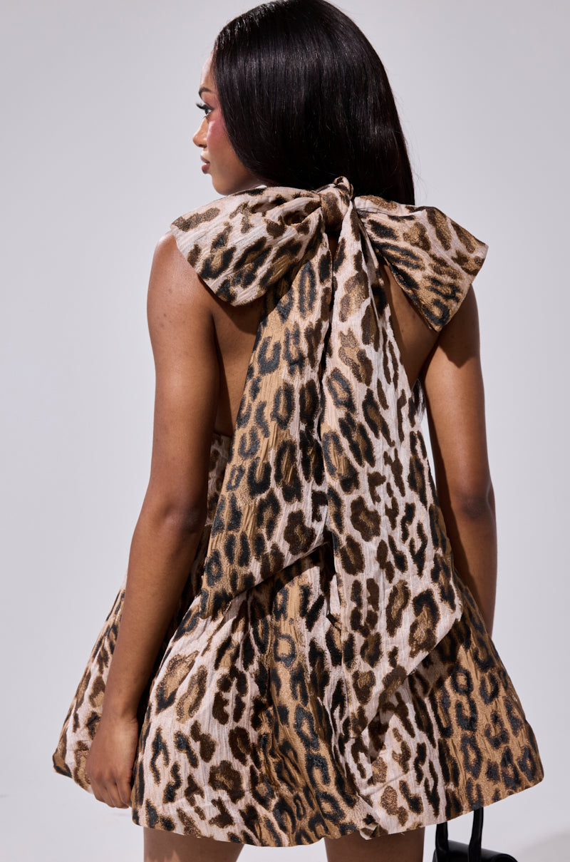 A woman with long dark hair models the PRINT OF THE SEASON BUBBLE MINI DRESS—a leopard print, sleeveless, backless mini dress with a large upper-back bow—standing with her back to a plain background.