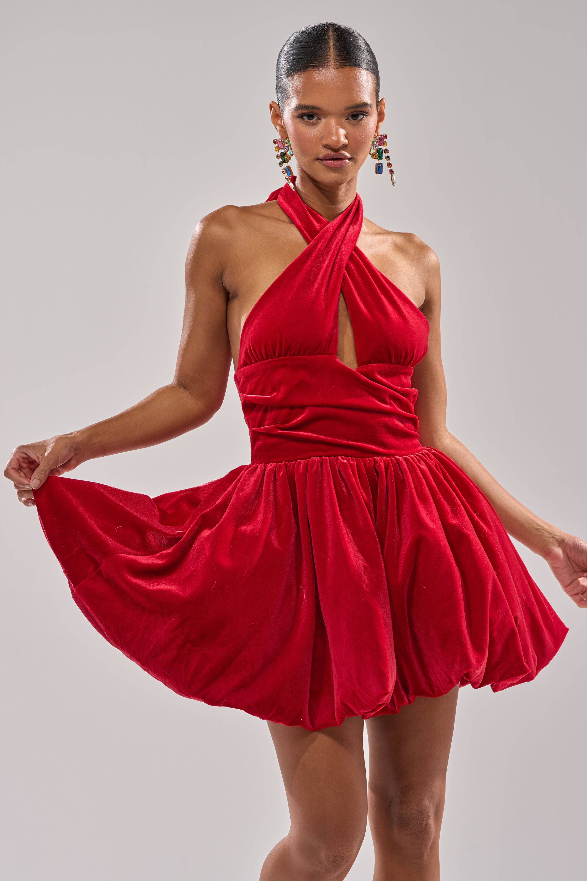 A woman models the RUBY ROSE VELVET MINI DRESS, featuring a sleeveless halter neckline and a dramatic bubble skirt. She pairs it with slicked-back hair and bold statement earrings while holding out the dress’s sides.