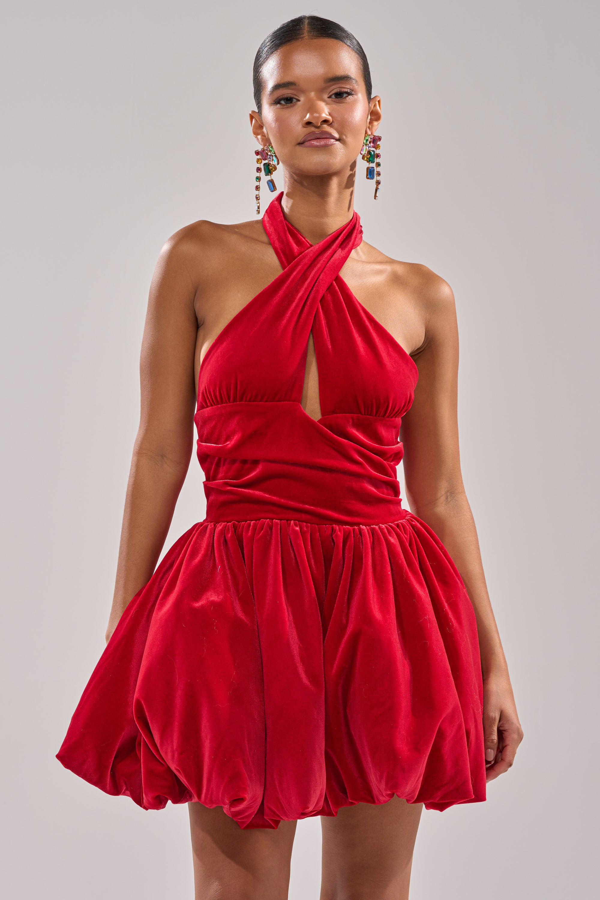 A woman models the RUBY ROSE VELVET MINI DRESS, featuring a vibrant red hue, cross-front halter neckline, and bubble skirt—ideal for holiday parties. She wears colorful drop earrings and has her hair slicked back against a light gray background.