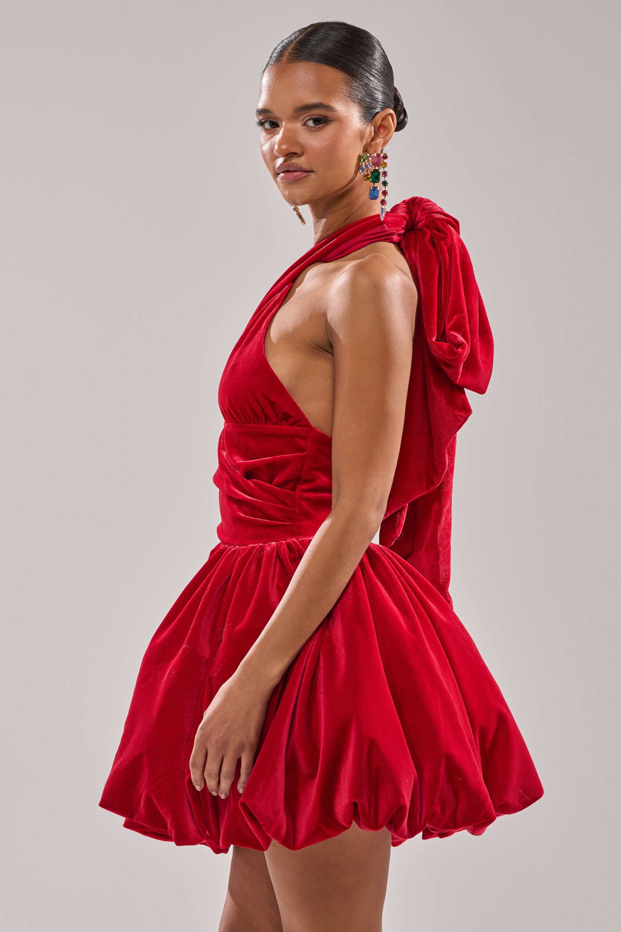 Wearing the RUBY ROSE VELVET MINI DRESS, featuring a bright red hue, voluminous bubble skirt, and a bold shoulder bow, she stands in profile against a light gray background—the perfect dress for any holiday party.