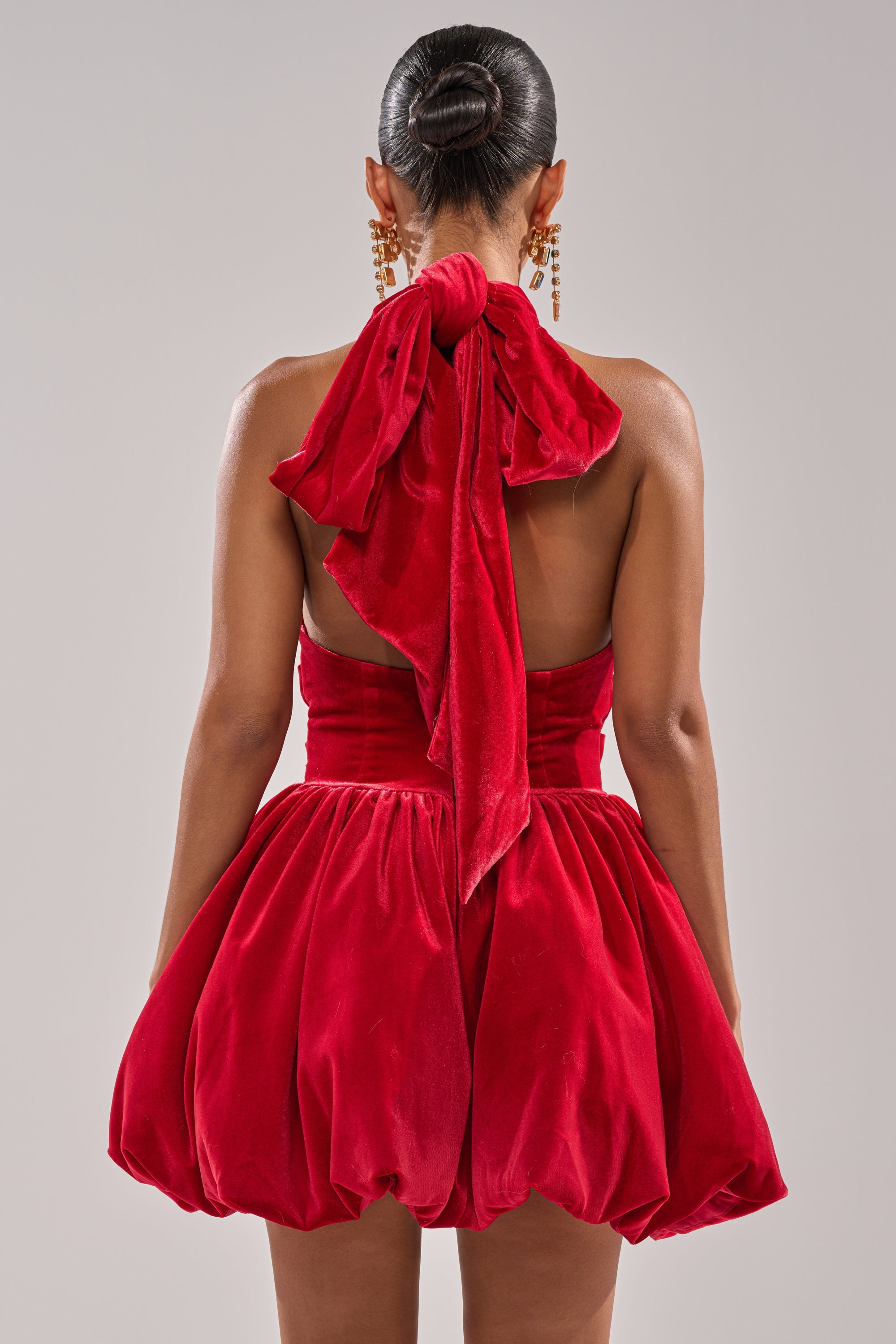 A woman with a sleek bun is seen from behind wearing the RUBY ROSE VELVET MINI DRESS—a red fit and flare style with a large neck bow, voluminous skirt, and dangling gold earrings.