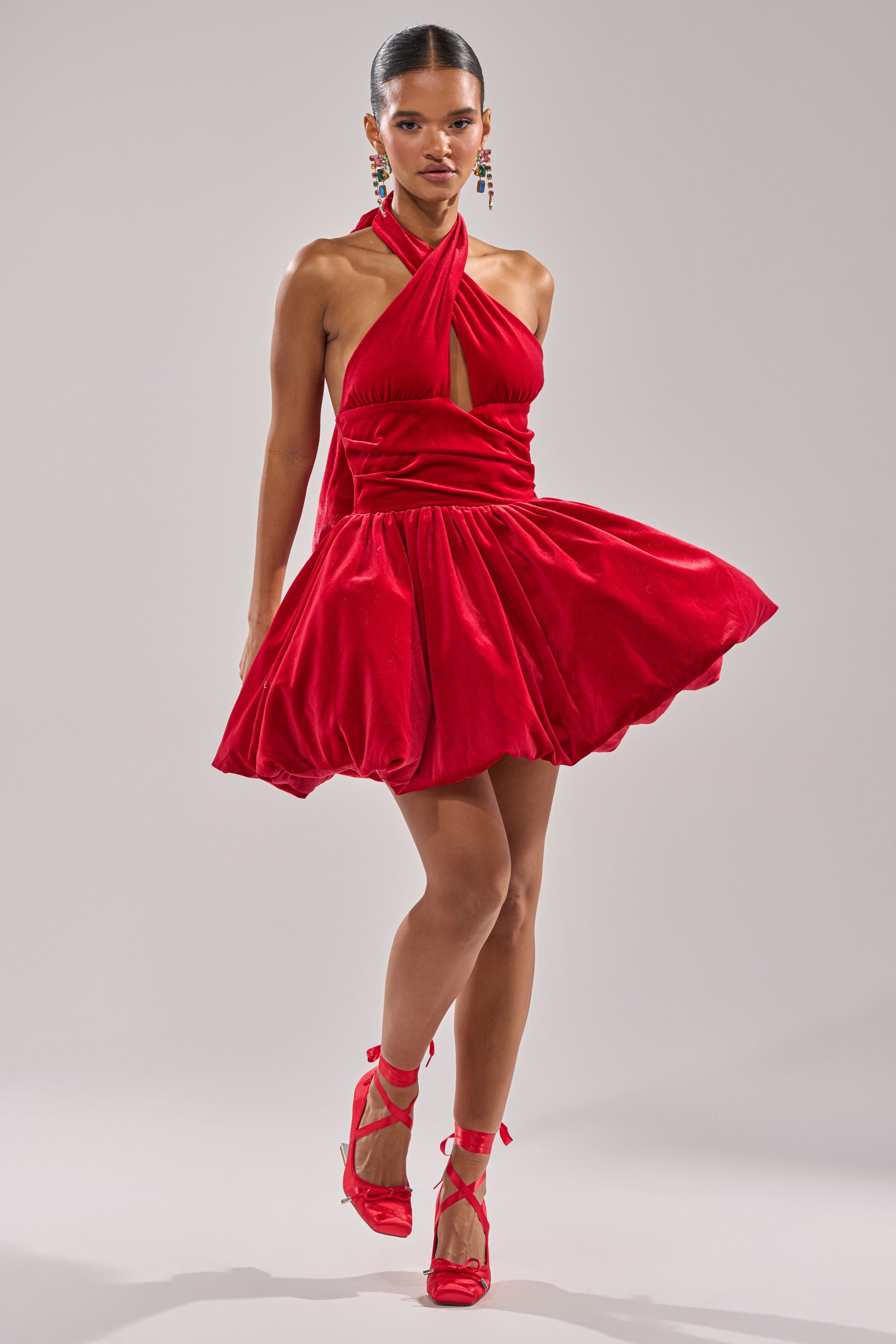 A woman poses confidently against a plain light background wearing the RUBY ROSE VELVET MINI DRESS, a sleeveless bright red fit-and-flare style with a halter neck and flared skirt, paired with matching red high heels.