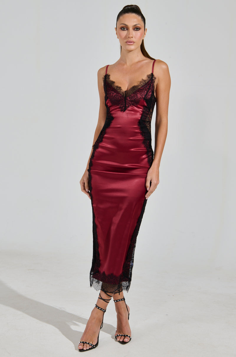 A model gracefully poses in the "SHOW US WHAT YOU GOT SATIN MIDI DRESS," a luxurious form-fitting red satin midi dress with black lace detailing and a V-neckline, standing elegantly in strappy heels against a plain backdrop.