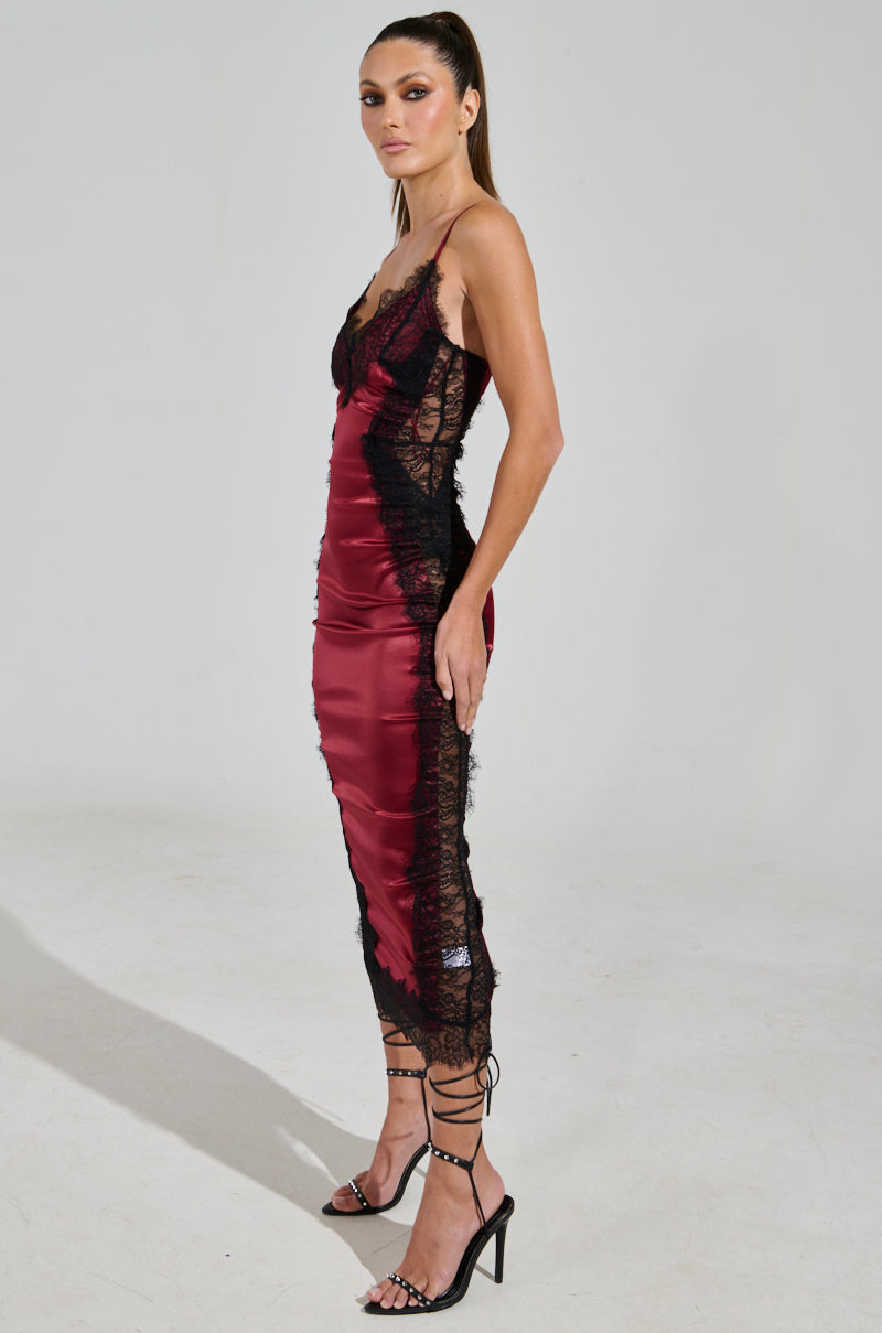 A woman poses side-on, highlighting the luxurious design of the fitted SHOW US WHAT YOU GOT SATIN MIDI DRESS with a sweetheart neckline in red and black lace. She stands against a plain background, wearing strappy black high-heeled sandals, her hair styled in a sleek ponytail.