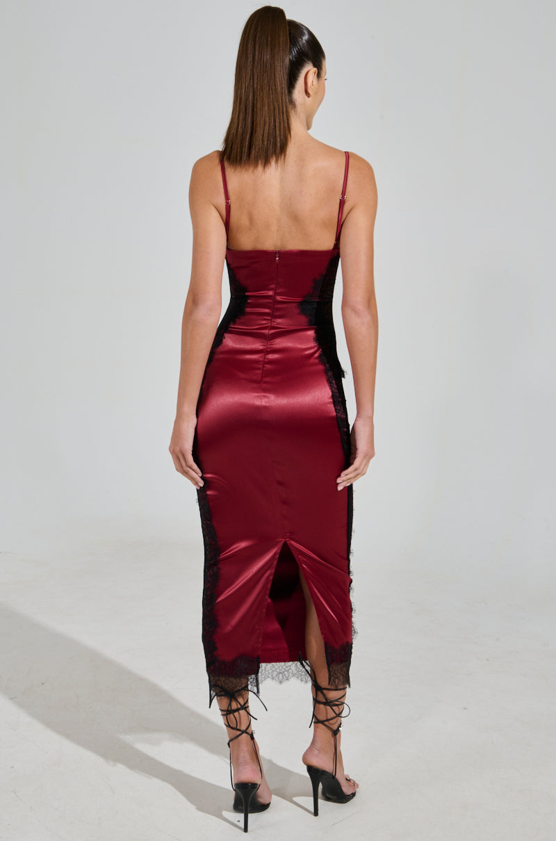 A woman with long, straight hair wears the SHOW US WHAT YOU GOT SATIN MIDI DRESS, a fitted burgundy satin midi dress adorned with black lace accents. With a sweetheart neckline, thin straps, and back slit, she stands against a plain background in black high-heeled sandals, exuding elegance.