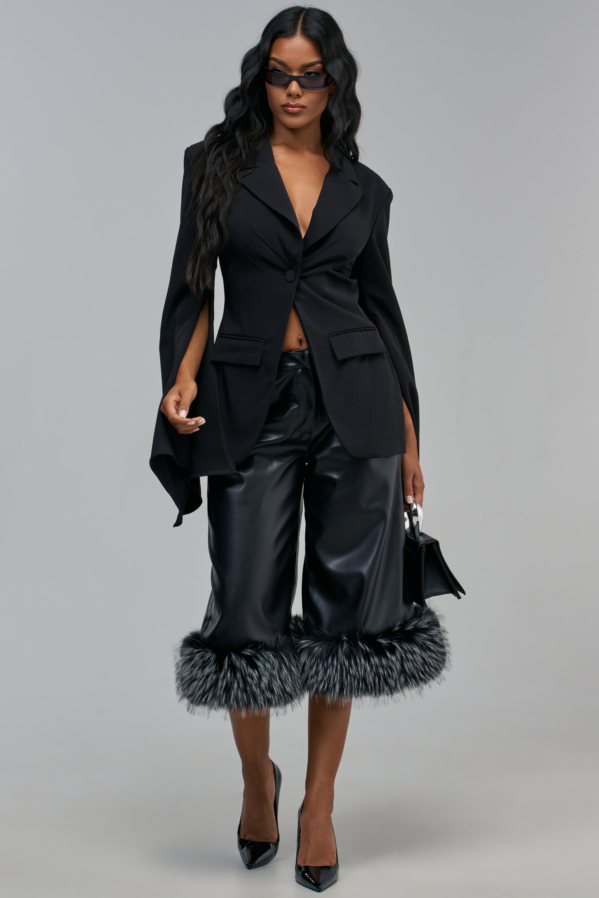 A woman wears a black blazer with slit sleeves and black faux leather MIND YOUR OWN FUR TRIM BERMUDA SHORTS with fur-trimmed hems, paired with black pumps and a small handbag.