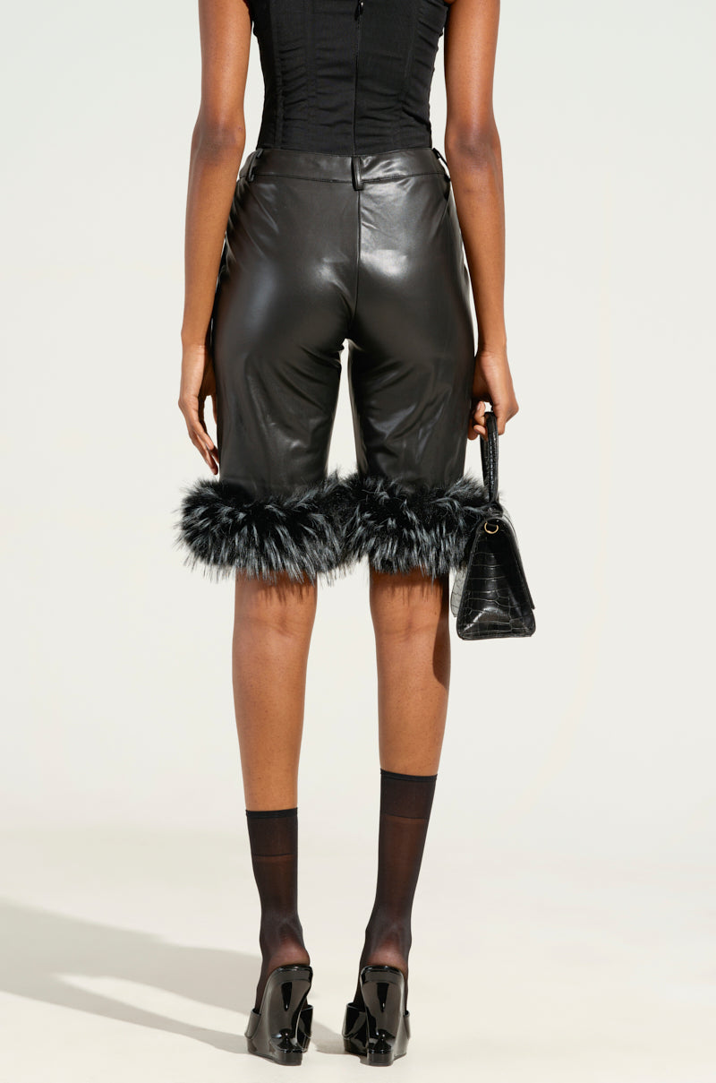 A person in the "Light As A Feather" black Bermuda shorts with dual-tone faux fur trim stands facing away, holding a black handbag. They wear a sleeveless top, sheer black socks, and black heels. The background is neutral.