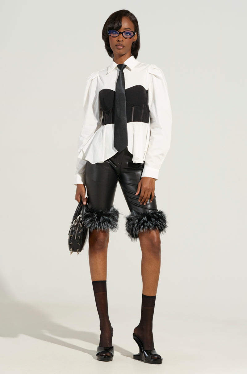 A fashion model showcases a white blouse with puffed sleeves, a black tie, and a corset. They highlight the LIGHT AS A FEATHER BLACK BERMUDA SHORTS featuring dual-tone faux fur trim at the knees. The ensemble is complemented by black knee-high socks, heels, and a small handbag. Finishing touches include short hair and glasses.