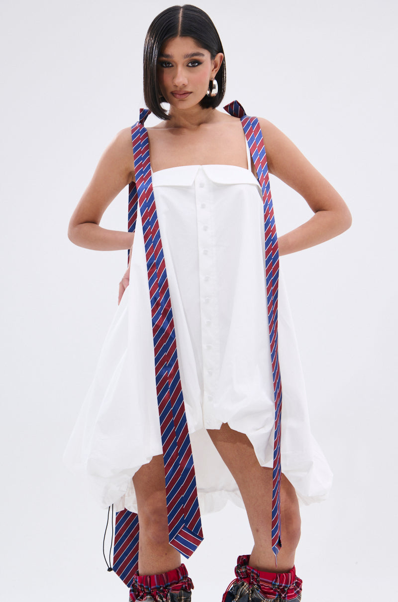 A woman models the OFFICE SIREN POPLIN MINI DRESS, a white button-down style with red and blue striped necktie shoulder straps. She pairs it with plaid leg coverings and has sleek, shoulder-length hair against a plain white background.