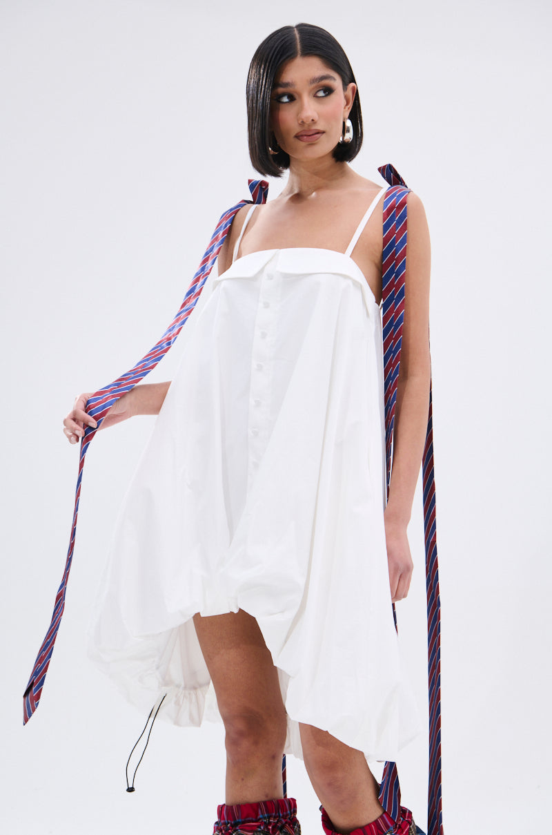 A woman showcases the OFFICE SIREN POPLIN MINI DRESS, a relaxed white button-down mini with adjustable tank straps. Her straight shoulder-length dark hair is adorned with vibrant striped ribbons cascading over her shoulders.