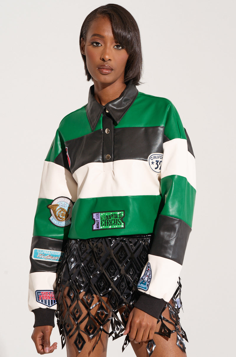 A person stands against a plain background, wearing the BIGGEST FAN LONG SLEEVE RUGBY TOP, which features green, white, and black striped color block design with embroidered letterman patches and a black collar. They also have on a black geometric skirt made of diamond-shaped pieces. Their hair is straight and shoulder-length.